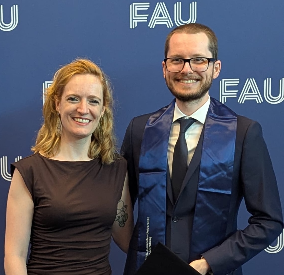 The image shows the author and his wife in front of a blue background that has branding by the FAU Erlangen-Nürnberg.
